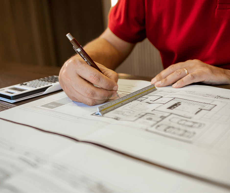 An architect sitting at a desk and using a rule on a blueprint. Next to them a calculator is present.