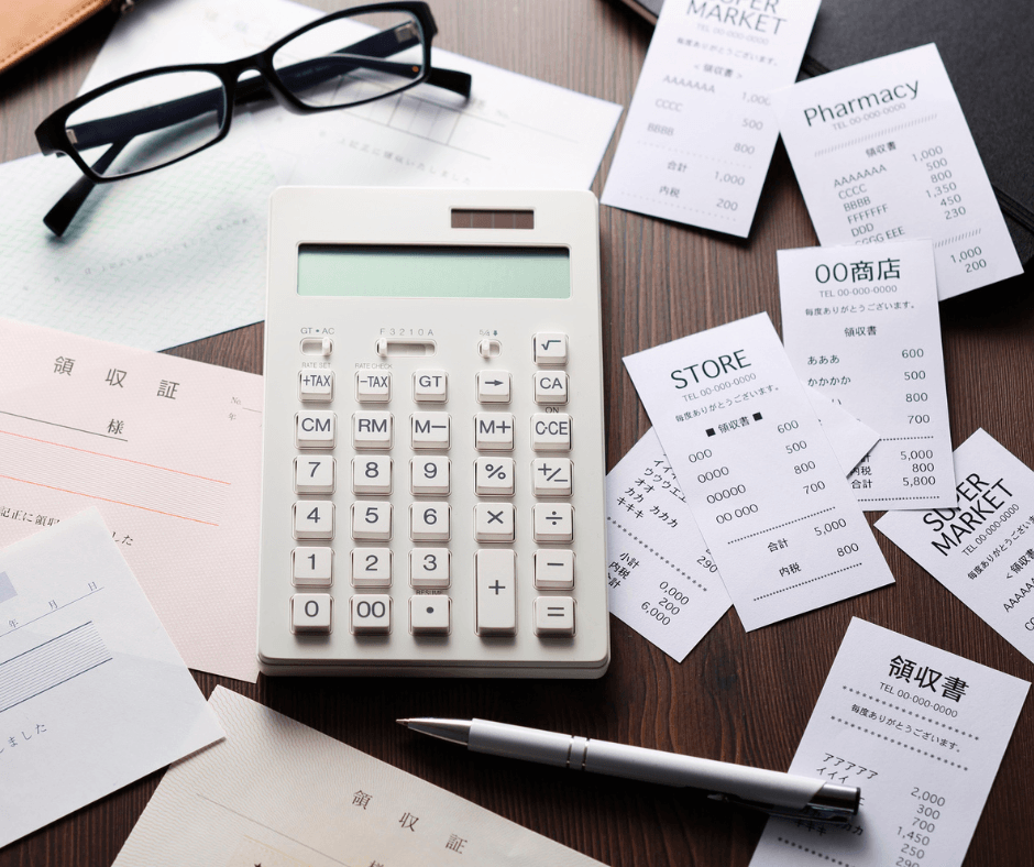 A calculator in the center surrounded by receipts. There are glasses above the math device and a pen below it. Demonstrates budget planning in every day.