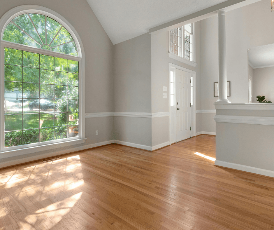An empty room with an arched window to the left. The floor is a light brown wood, and the walls are a soft gray. The right has an entry way into the room.