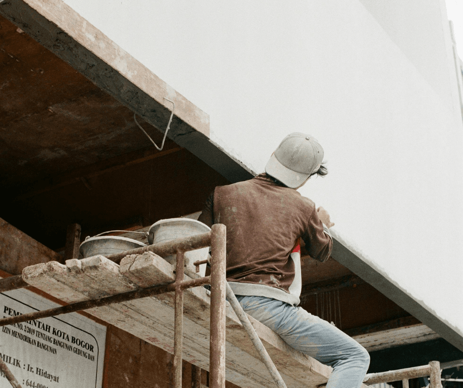 Someone on a scaffolding painting something up high. Their back is facing you in the picture, with their leg over scaffold's platform.