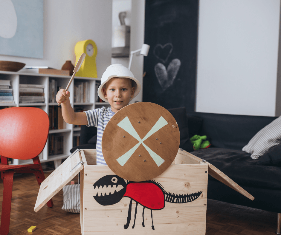 A child with a construction hat in a box playing pretend. He has a spatula sword and a fake shield to protect himself.