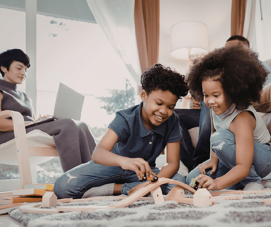 Two children playing with trains in front of their parents. They are smiling while a background person (assumedly the mom) is on the computer.