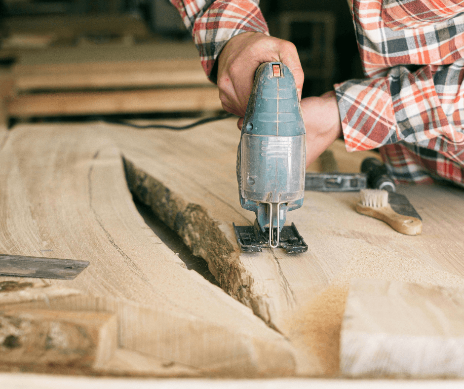 A picture that shows a carpenter carving wood away from a split piece of wood. It looks like the person is creating a table, with the carving tool being held by two hands. Loose wooden material is shown underneath a gap in the wood.